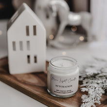 Kép betöltése a galériamegjelenítőbe: Nutcracker szójagyertya ámbra üvegben, nyitott állapotban, mellette havas ág és porcelán házikó, karácsonyi háttérfénnyel.