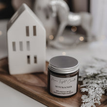 Kép betöltése a galériamegjelenítőbe: Nutcracker szójagyertya nyitott tetővel, fa tálcán, ünnepi hangulatú dekorációval és bokeh fényekkel.