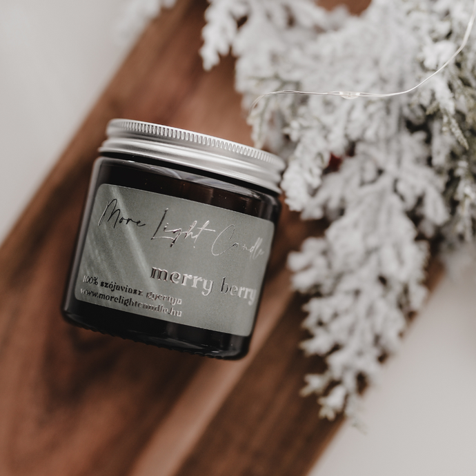 Merry Berry szójagyertya fa tálcán, friss szilvával és vaníliarúddal körülvéve, ünnepi hangulatú háttérrel.