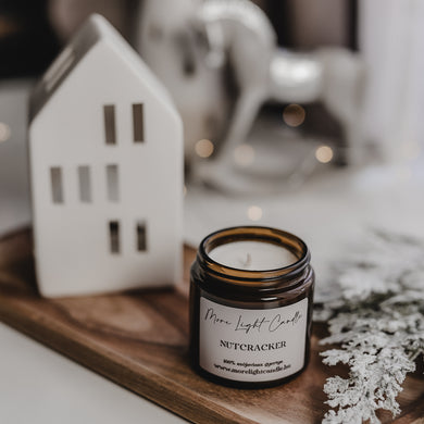 Nutcracker szójagyertya ezüst tetővel, fa tálcán, háttérben porcelán házikó és karácsonyi fények.